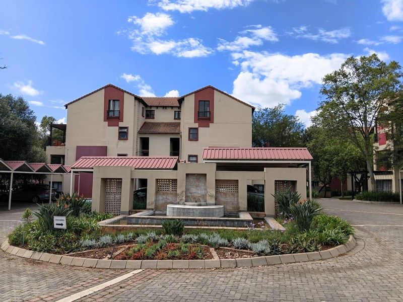 Front exterior view of the apartment complex featuring a central water fountain, landscaped garden beds, and covered parking structures.