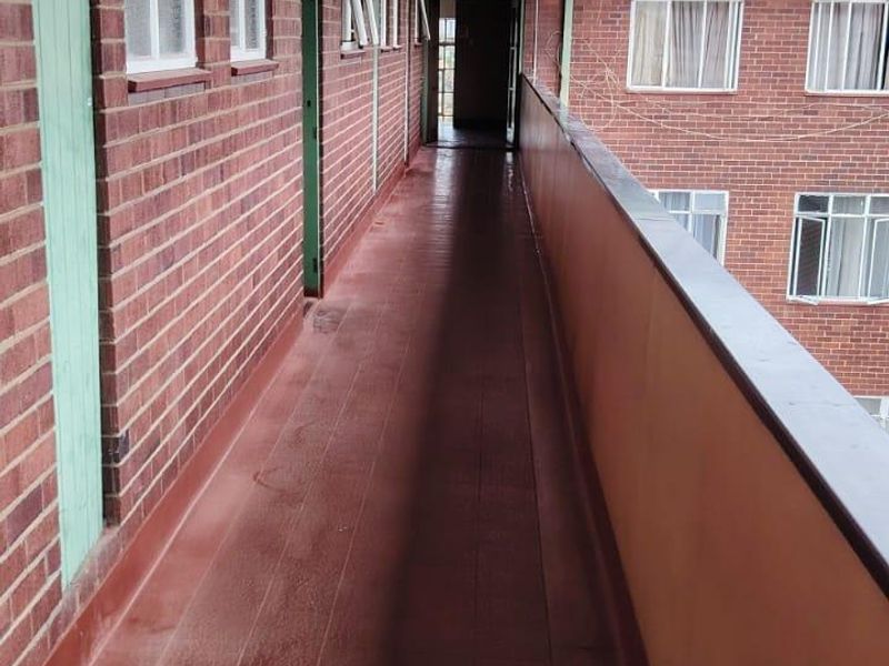 An exterior apartment access walkway featuring brick walls, tiled flooring, and a metal balustrade, showing signs of wear on the ceiling and floor surfaces.