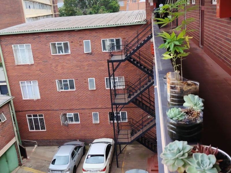 View of the apartment building exterior, featuring a brick facade, external metal staircase, and a tiled walkway overlooking the parking area.