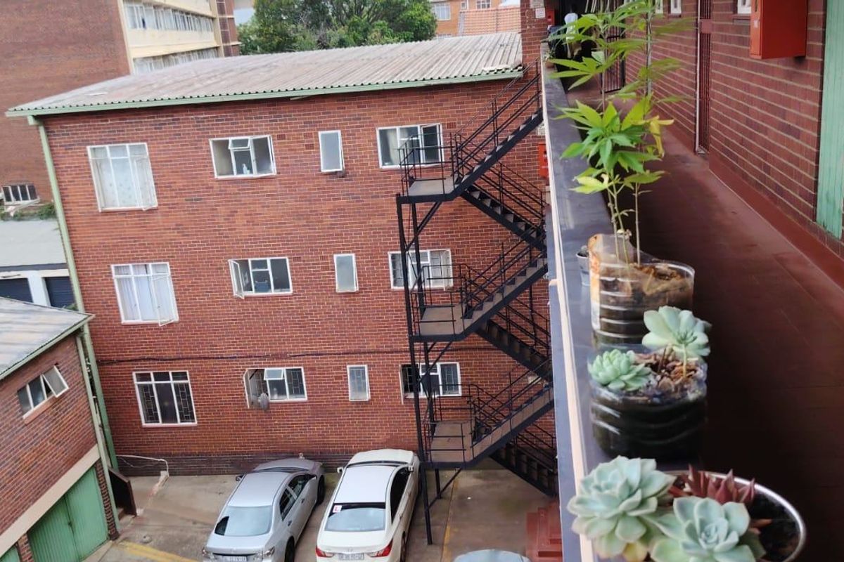 View of the apartment building exterior, featuring a brick facade, external metal staircase, and a tiled walkway overlooking the parking area.