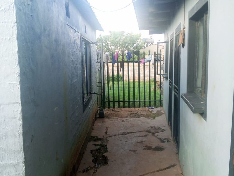 A narrow exterior side passage between two building structures featuring a concrete walkway and a metal security gate leading to the rear garden area.
