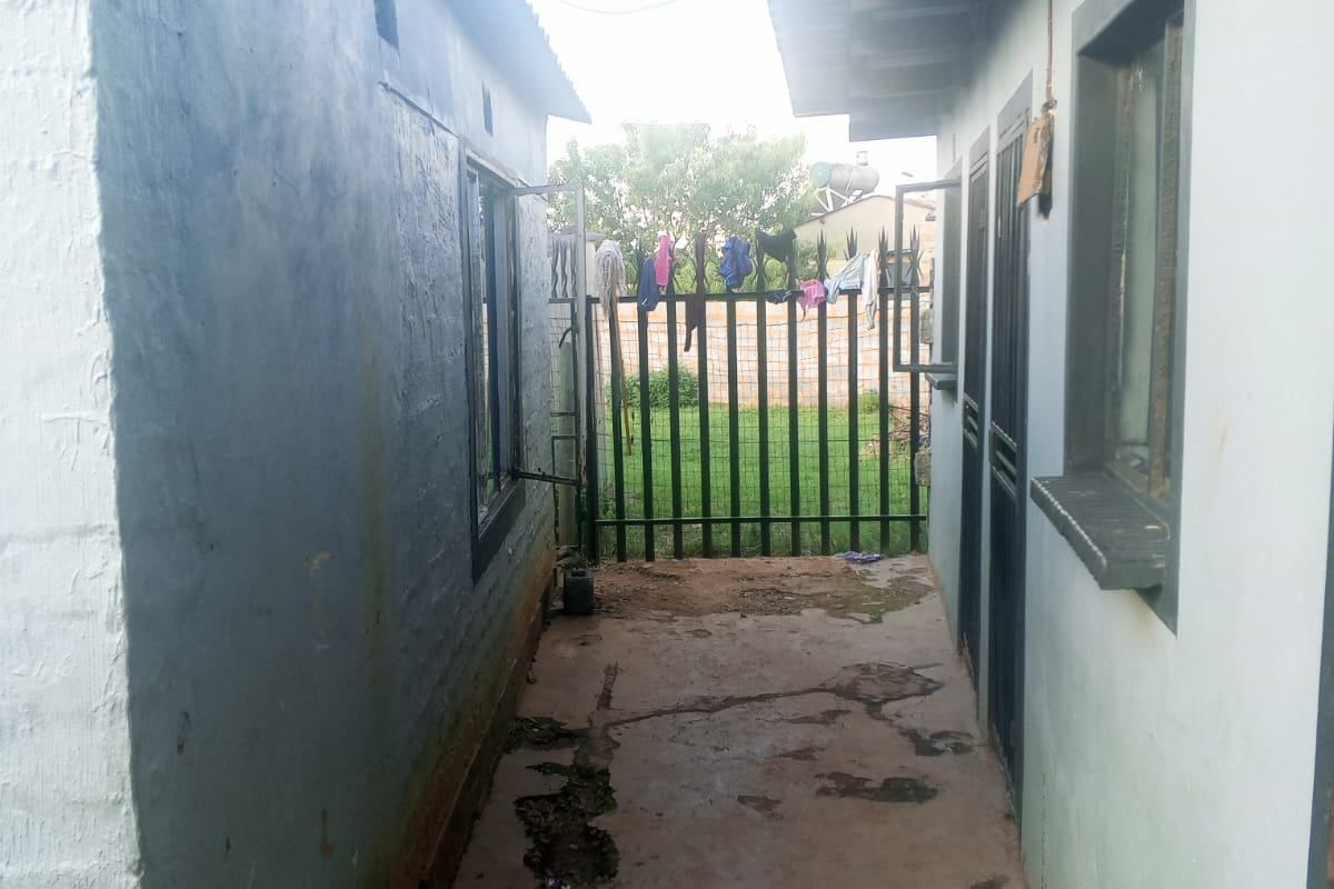 A narrow exterior side passage between two building structures featuring a concrete walkway and a metal security gate leading to the rear garden area.