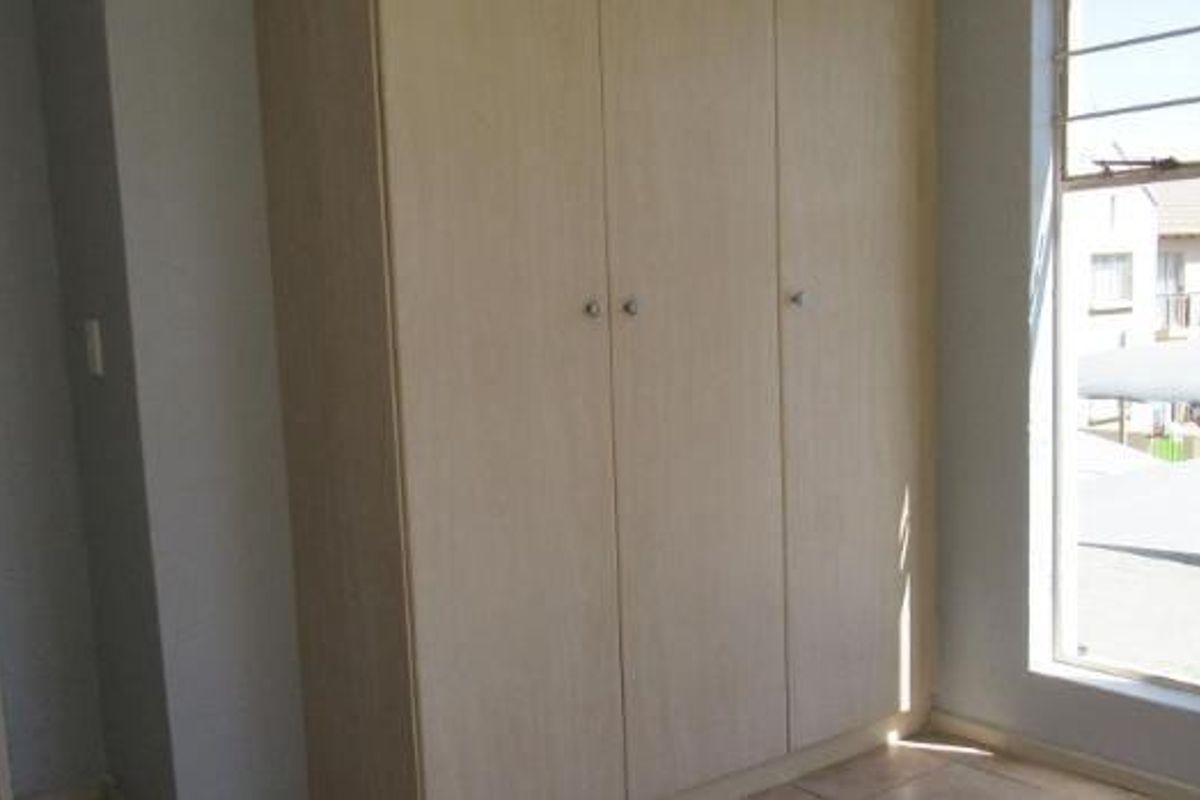 A bright, empty bedroom featuring built-in wooden wardrobes, tiled flooring, and a window with security bars.