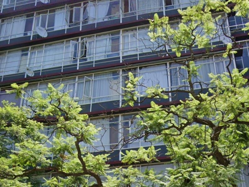 A multi-story apartment building facade featuring large windows and balcony enclosures, viewed from street level with mature trees in the foreground.