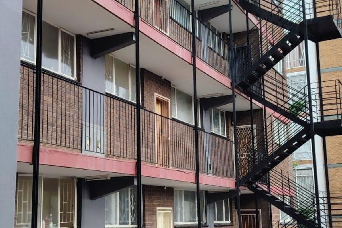 Exterior view of a multi-story apartment building featuring a prominent external steel fire escape staircase and brick facade with balcony access.