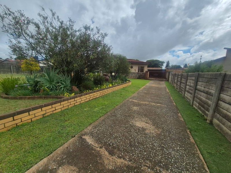 A front exterior view featuring a long concrete driveway, a brick-edged garden bed, and a secure metal gate leading to the property entrance.