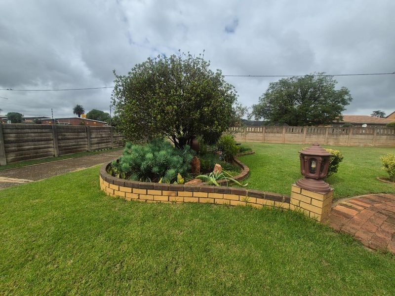A well-maintained garden area featuring a curved brick retaining wall, established shrubs, and a paved pathway enclosed by a concrete boundary wall.