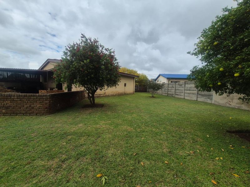 A spacious rear garden featuring a well-maintained lawn, mature trees, a brick boundary wall, and a covered patio area.