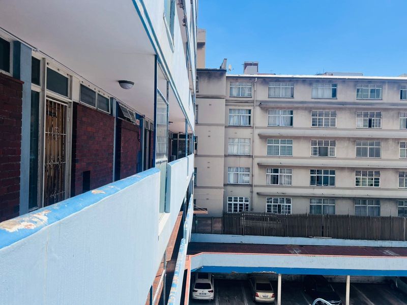 View of the apartment building exterior, featuring a communal walkway, balcony balustrades, and an adjacent parking area.