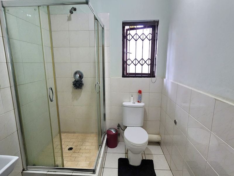 A functional bathroom featuring a glass-enclosed shower, standard toilet, and tiled finishes throughout, complete with a window featuring a security gate.