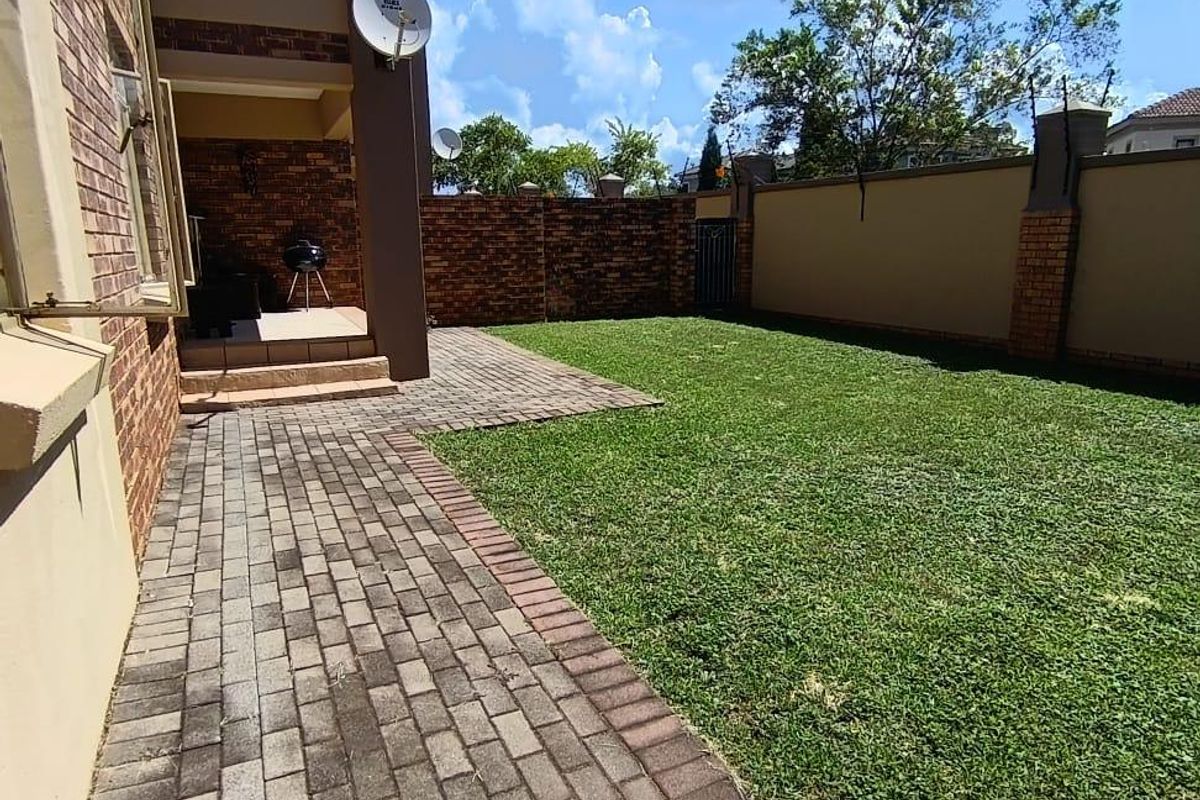 A private rear garden featuring a paved walkway, a well-maintained lawn, and a covered patio area enclosed by a perimeter brick wall.