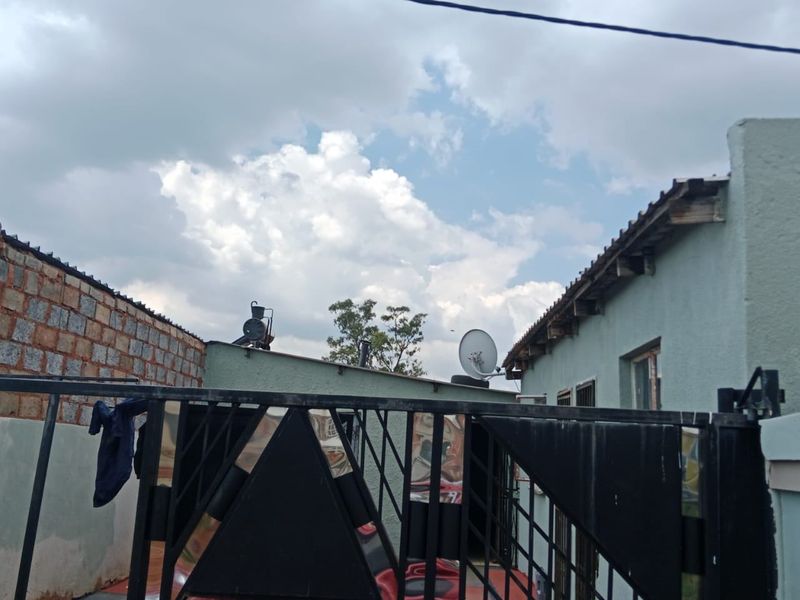 Front entrance view featuring a metal security gate, tiled driveway area, and brick boundary wall under a cloudy sky.