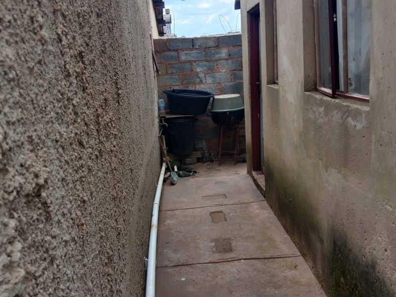 A narrow side passage or rear exterior area featuring concrete paving, exposed brick boundary wall, and external plumbing fixtures.