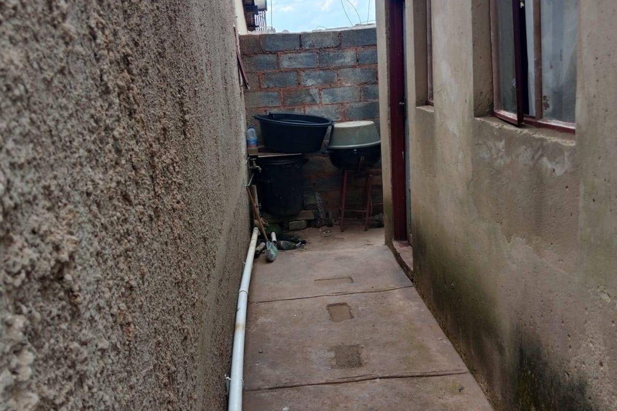 A narrow side passage or rear exterior area featuring concrete paving, exposed brick boundary wall, and external plumbing fixtures.