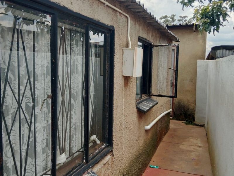 A narrow rear exterior pathway showing the side of the house with security-barred windows, corrugated roofing, and visible signs of exterior wall weathering.