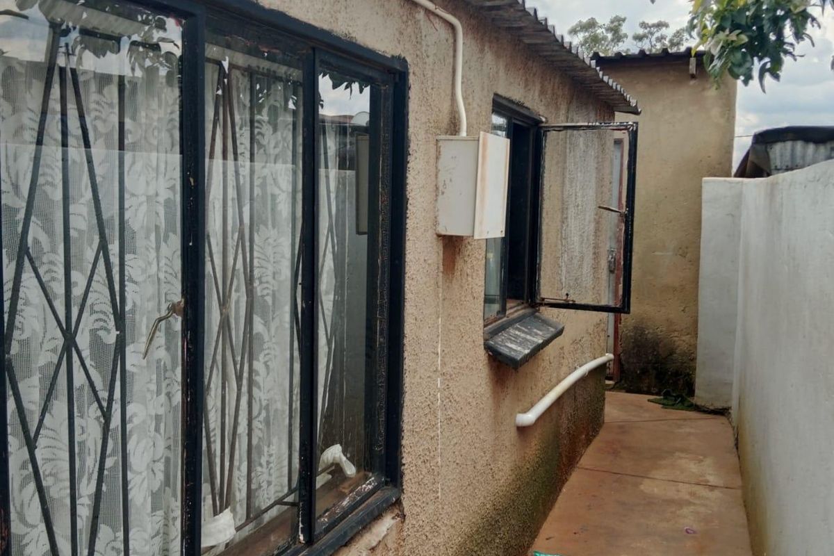 A narrow rear exterior pathway showing the side of the house with security-barred windows, corrugated roofing, and visible signs of exterior wall weathering.