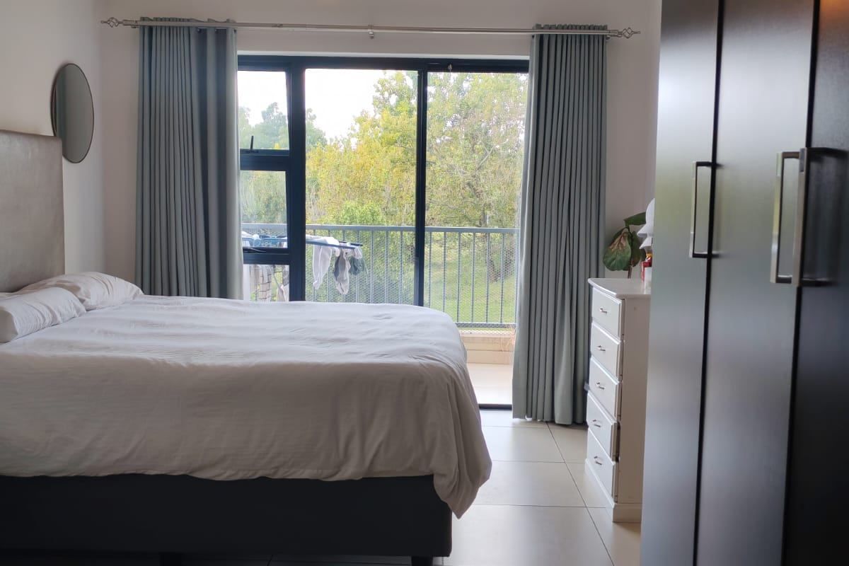 A well-lit bedroom featuring large sliding glass doors leading to a balcony, neutral tiled flooring, and a spacious built-in wardrobe.