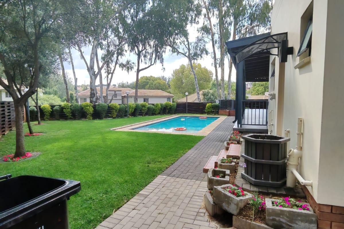 A well-maintained rear garden featuring a swimming pool, paved pathway, and covered patio area, surrounded by established trees and landscaping.