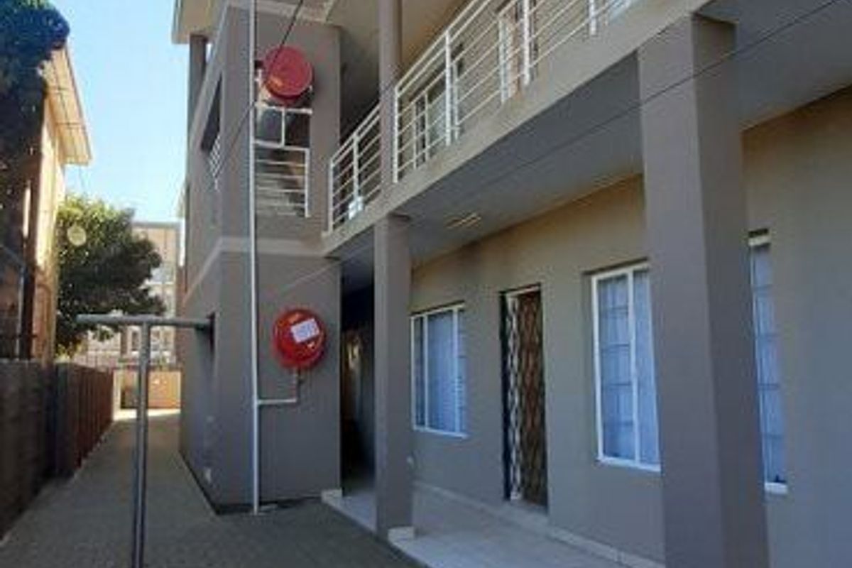 Exterior view of an apartment building featuring multi-level balconies with metal railings, paved ground level walkway, and external fire safety equipment.