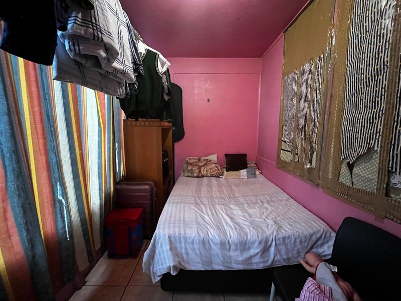 A compact bedroom featuring tiled flooring and pink painted walls, currently used as a multi-purpose living and storage space.