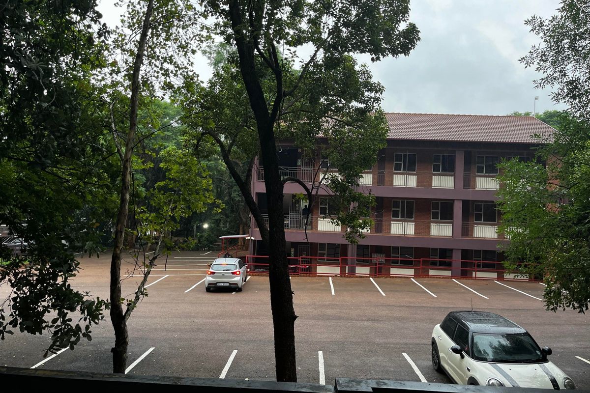 View of the apartment building exterior featuring a brick facade, tiled roof, and a paved parking area.