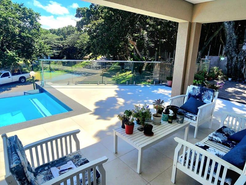A spacious covered patio featuring modern tiled flooring and a glass balustrade, overlooking a private swimming pool and lush garden area.