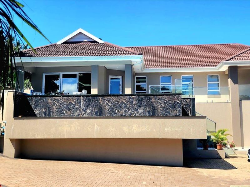 A modern multi-level residential property featuring a paved driveway, stone-clad retaining wall, glass balustrades, and a tiled roof.
