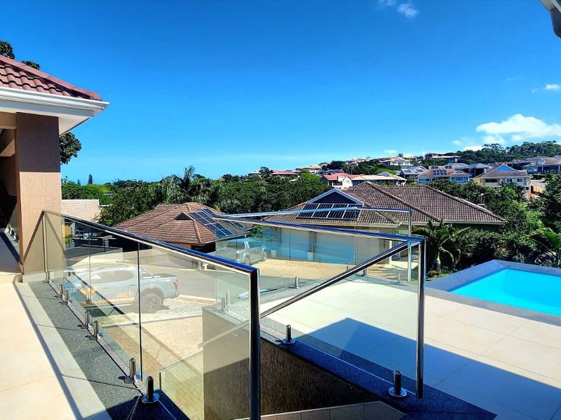Spacious covered balcony featuring modern tiled flooring and a sleek glass balustrade, overlooking a private swimming pool and surrounding residential landscape.