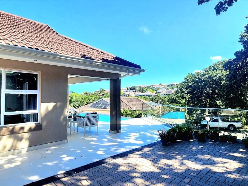 Spacious covered patio featuring tiled flooring and a glass balustrade, overlooking a swimming pool and landscaped garden area.