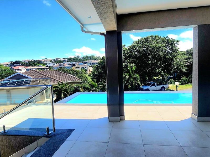 A spacious covered patio overlooking a swimming pool with glass balustrades and expansive views of the surrounding residential area.