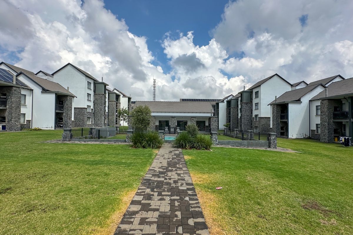 A view of the apartment complex grounds featuring a paved walkway leading through a well-maintained lawn towards the central communal area.