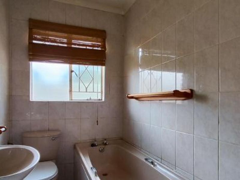 A functional bathroom featuring a bathtub, pedestal basin, and toilet with full-height wall tiling and security bars on the window.