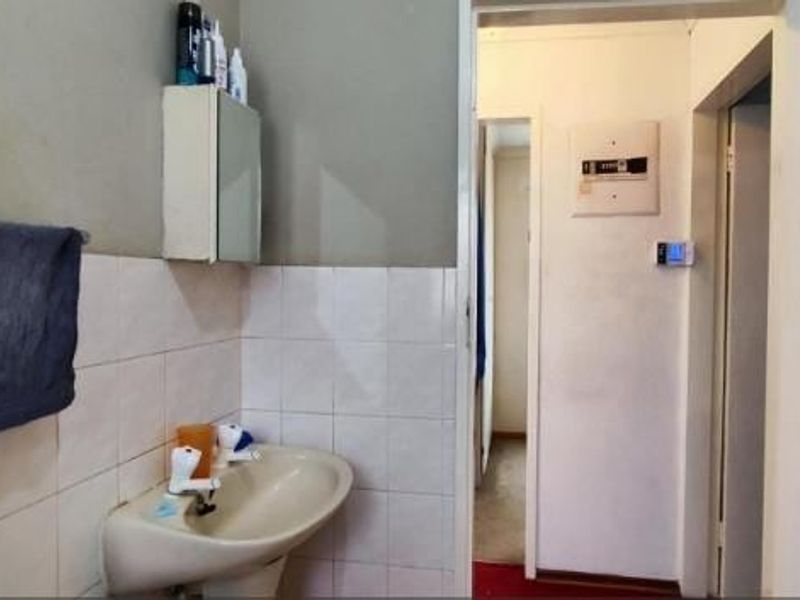 A functional bathroom featuring a pedestal washbasin, wall-mounted medicine cabinet, and partial ceramic wall tiling, leading into a hallway with an electrical distribution board.