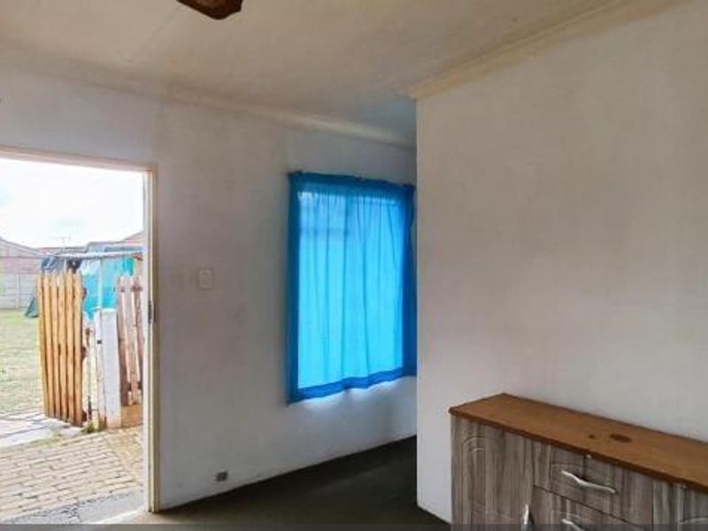 A sparsely furnished living area featuring a timber door leading to the exterior, a ceiling fan, and a window with blue curtains.