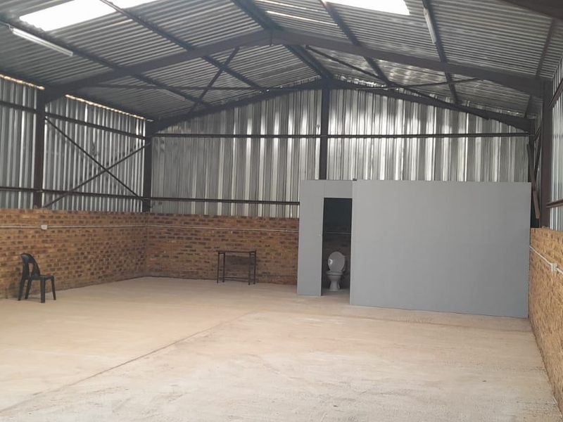 Spacious industrial utility area featuring high ceilings with exposed steel trusses, face brick walls, concrete flooring, and an integrated private toilet cubicle.