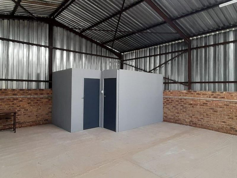 Industrial utility space featuring exposed brick walls, corrugated metal roofing, steel trusses, and internal partitioned office cubicles on a concrete floor.
