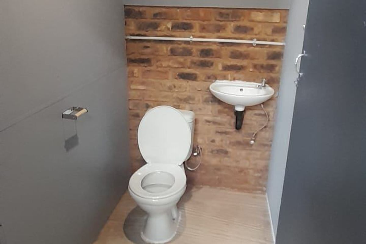 A compact industrial-style bathroom featuring an exposed brick wall, wall-mounted basin, and standard toilet, requiring finishing touches to the flooring and walls.