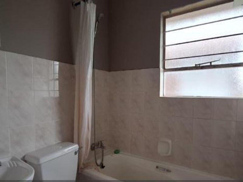 A functional bathroom featuring a bathtub with shower attachment, pedestal basin, and toilet, finished with neutral wall tiling and a window for natural light.