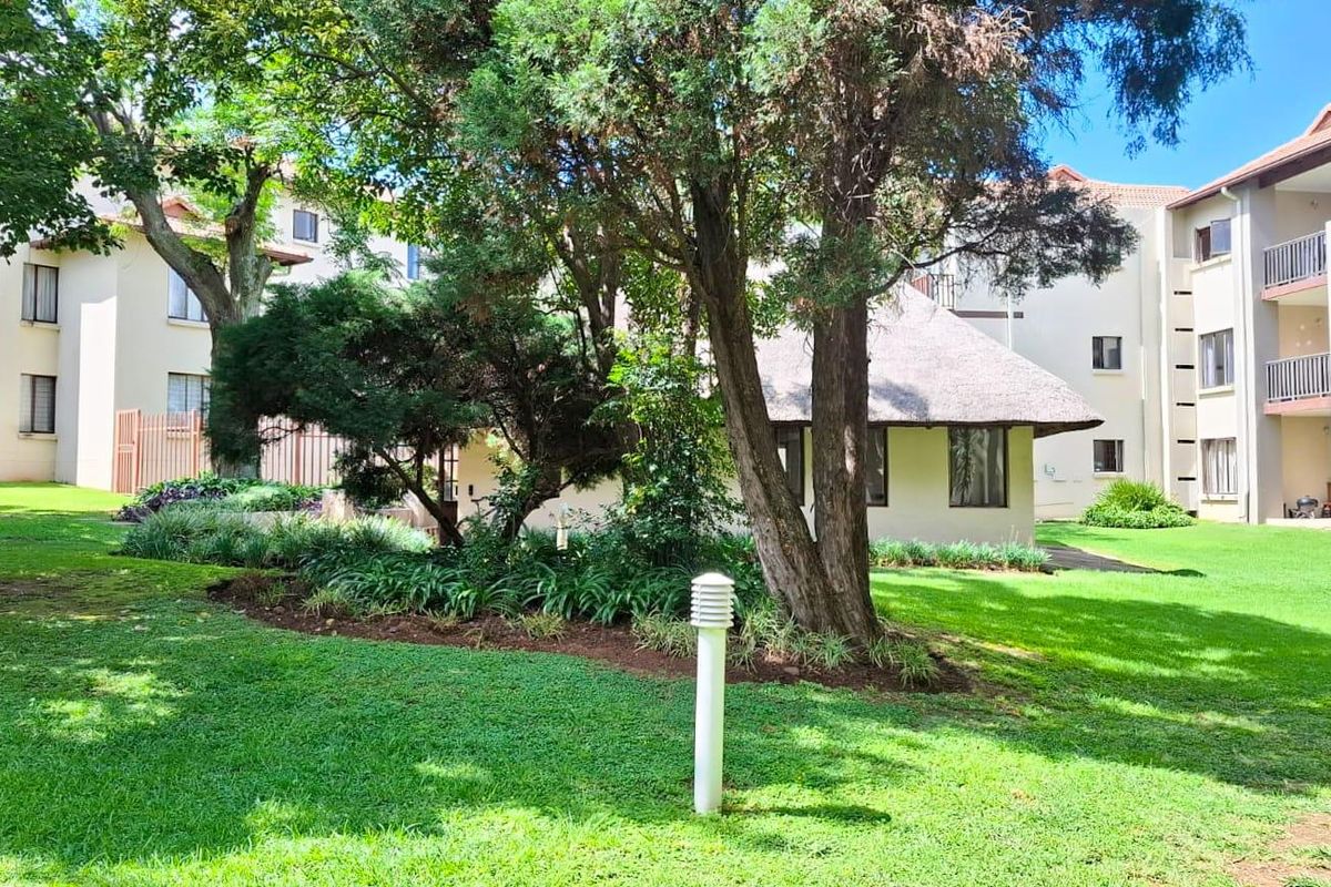 A well-maintained communal garden area featuring mature trees, landscaped beds, and a thatched-roof structure, surrounded by apartment building facades with balconies.