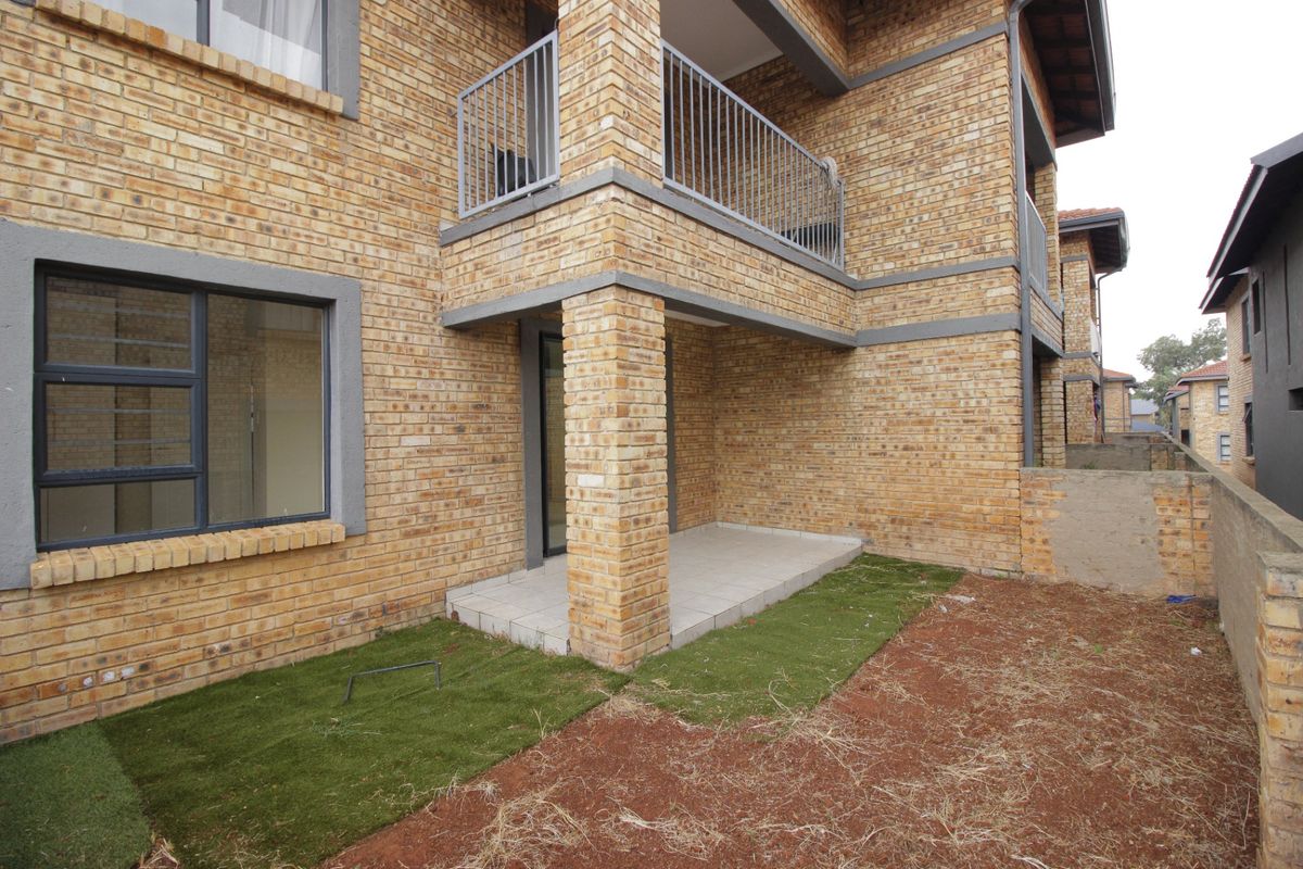 Rear view of a face brick apartment building featuring a ground floor tiled patio, upper balcony with metal railing, and an unfinished garden area.