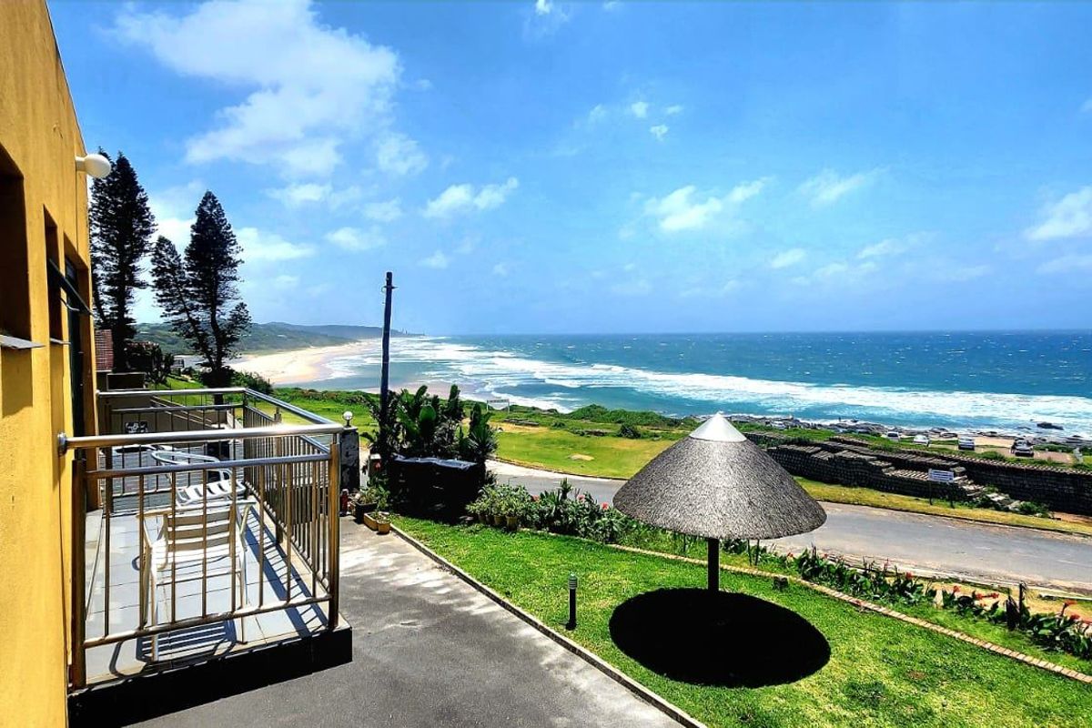 A rear exterior view featuring a balcony with stainless steel railings, a paved patio area, a thatched umbrella, and expansive ocean views.