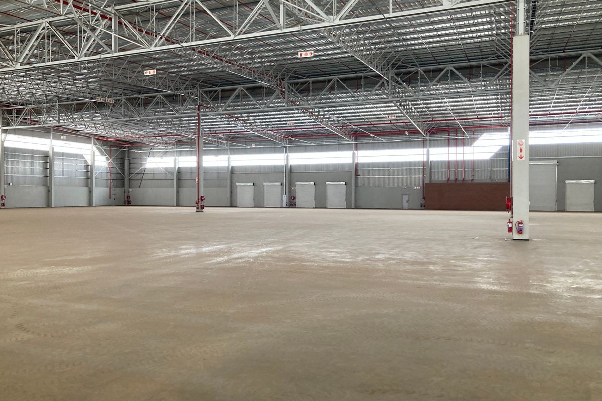 Spacious industrial warehouse featuring high ceilings with exposed steel trusses, concrete flooring, multiple roller shutter doors, and an integrated fire sprinkler system.