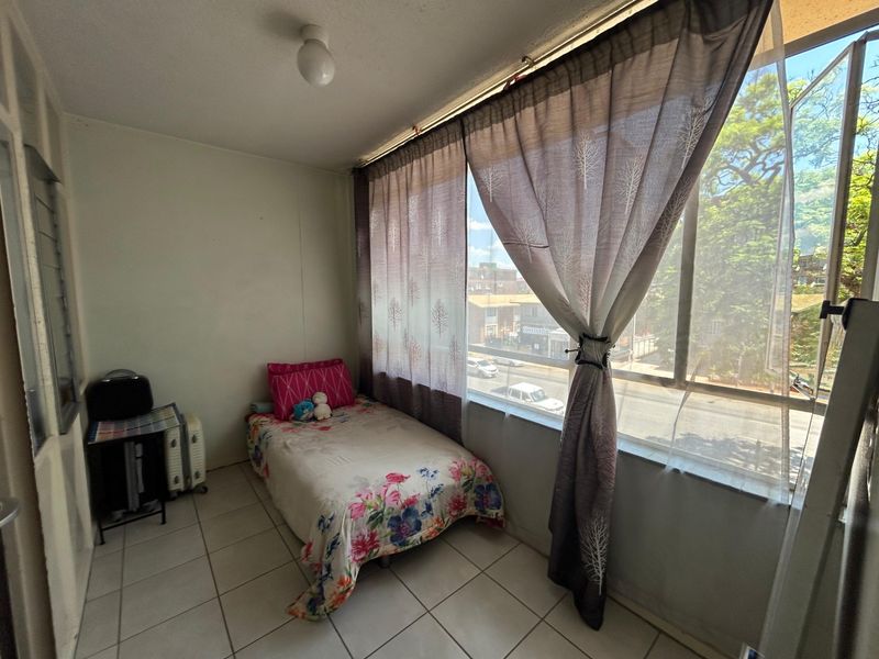 A compact bedroom featuring tiled flooring and a large window providing natural light and street views.