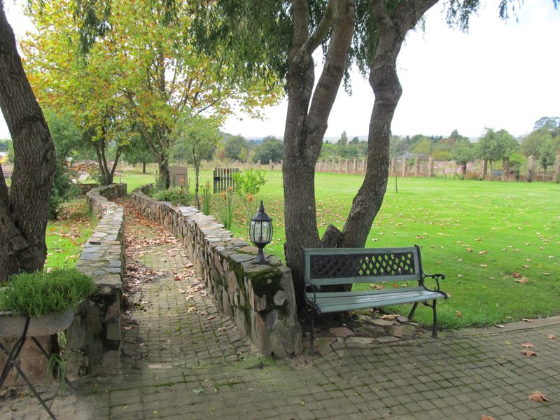 A spacious, well-maintained garden featuring a stone-walled walkway, mature trees, and a large lawn area enclosed by a brick perimeter wall.