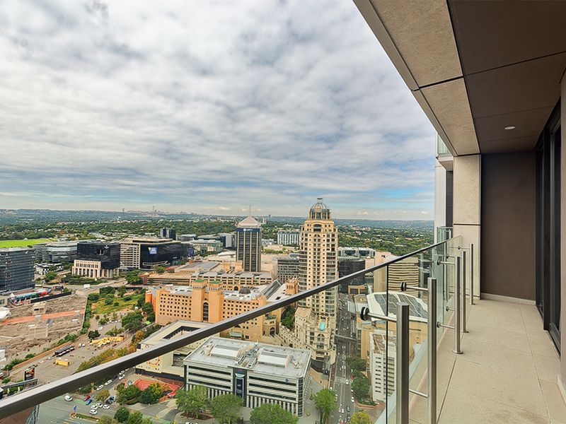 A modern apartment balcony featuring a glass balustrade and tiled flooring, offering expansive elevated views of the surrounding city skyline.