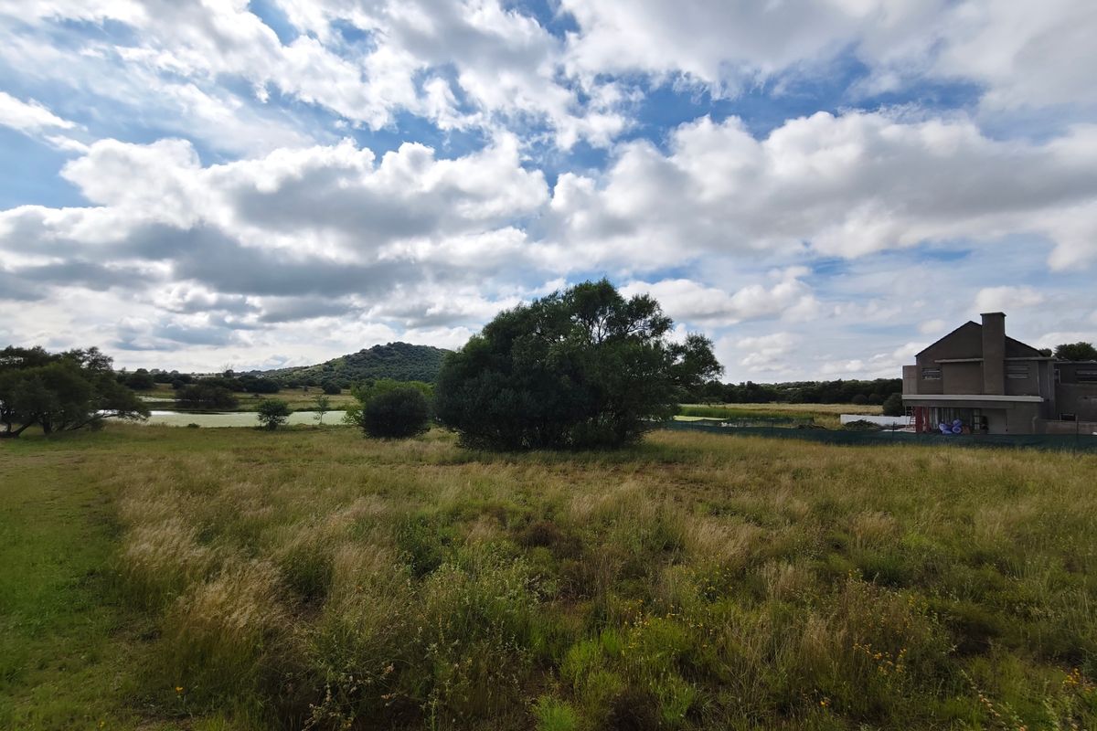 A scenic view of an expansive vacant residential plot featuring natural grassland, mature trees, and distant hills under a cloudy sky.