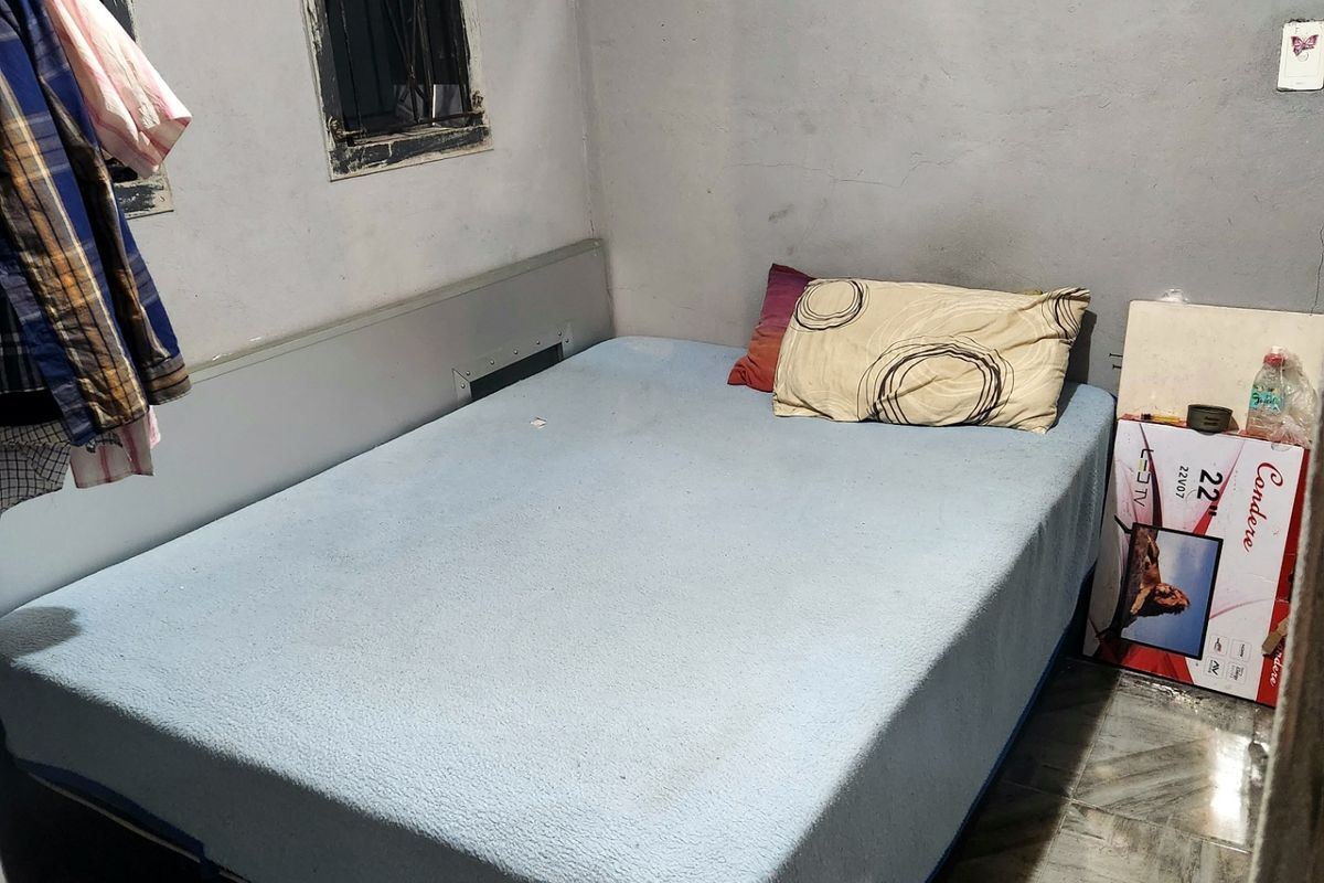 A compact bedroom featuring tiled flooring and windows with security bars, currently requiring cosmetic updates and wall repairs to improve the overall condition.