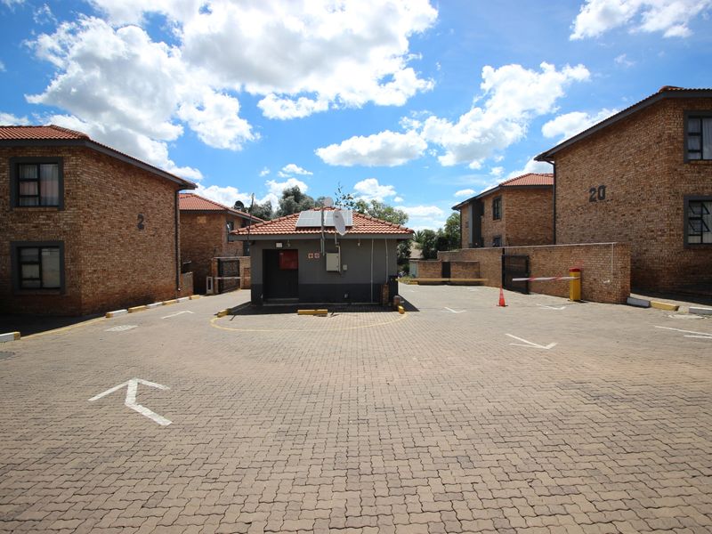 Exterior view of the apartment complex entrance featuring brick buildings, a paved driveway, and a central security guard house with solar panels.
