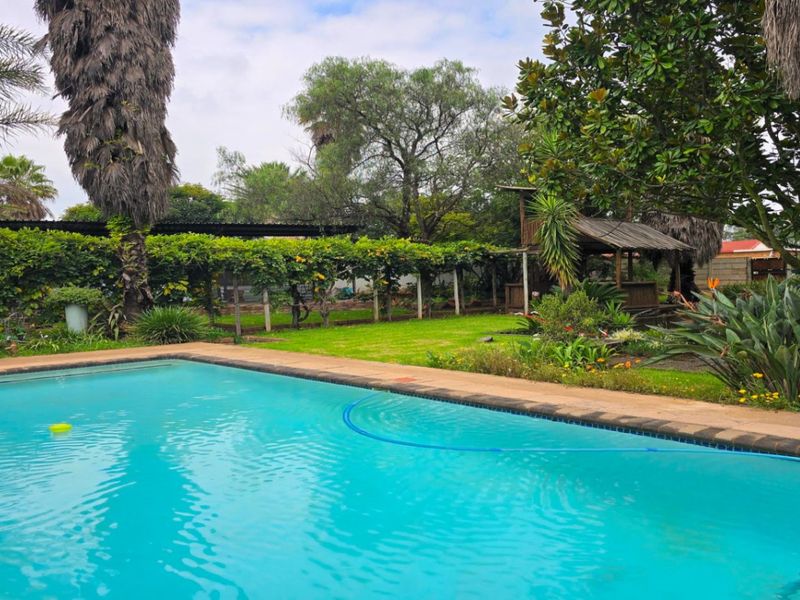 A spacious outdoor pool area featuring a swimming pool with tiled coping, surrounded by a lush garden, mature trees, and a shaded pergola structure.