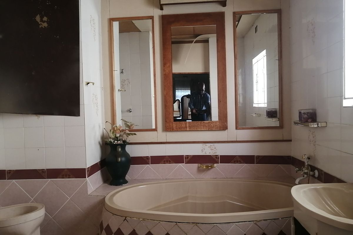 A dated bathroom featuring a built-in corner bathtub, pedestal sink, and wall-mounted mirrors with visible wear on the tiled surfaces.